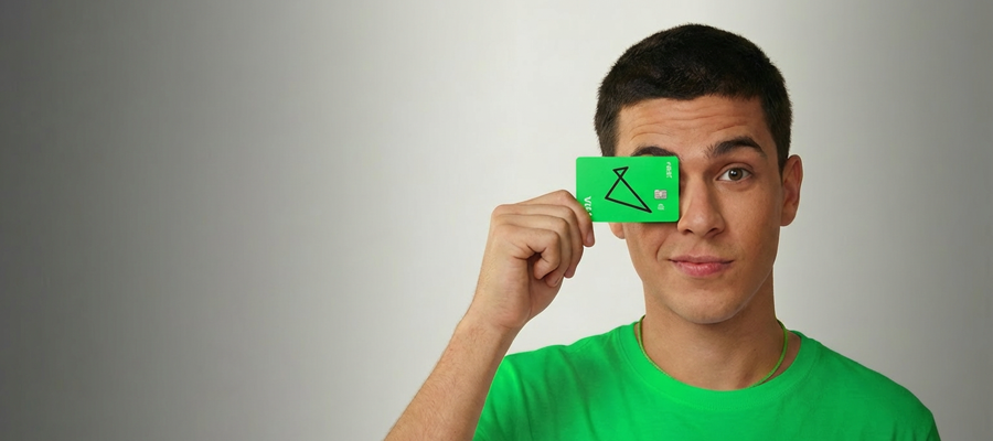 Uma mulher preta, com cabelos castanhos e ondulados, lisos e longos, segura na frente do olho esquerdo dela o cartão de crédito da cor verde neon do banco next. Ela veste uma blusa regata e um colar fino, ambos da cor verde neon.